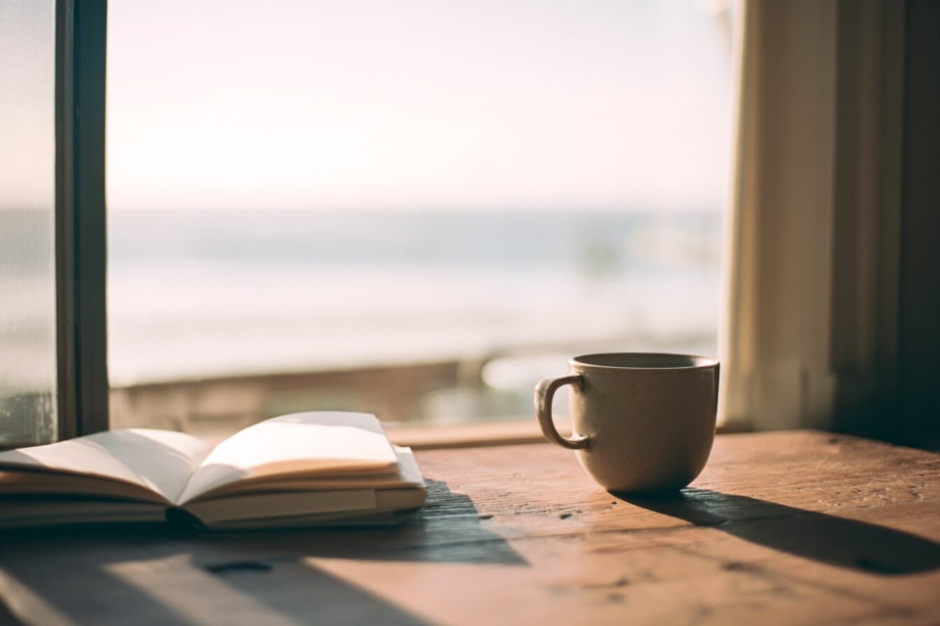 Morning light on a wooden desk with a notebook and a cup, beside a window looking out to sea.